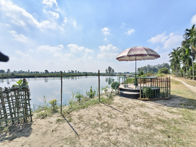 ​A peaceful, rustic seating area under a patterned umbrella, perched on the bank of a calm body of water under a bright, cloudy sky.